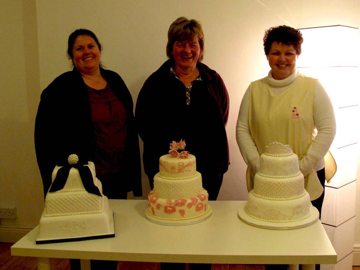 thre smiling woemnt with cakes in front of them
